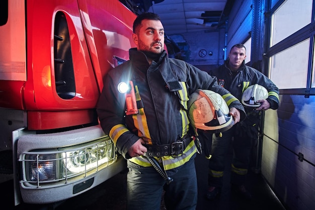 Kostenloses Foto zwei feuerwehrmänner in schutzuniform stehen neben einem feuerwehrauto in einer garage einer feuerwehr.