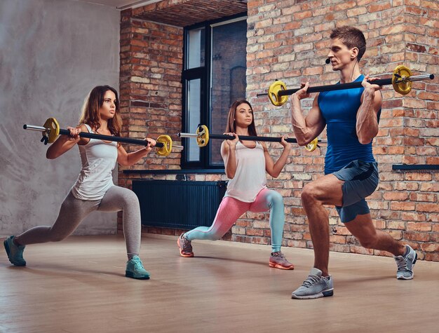 Zwei Fitnessfrauen und ein Trainer dehnen Kniebeugen mit Langhanteln in einem Club mit Loft-Interieur.