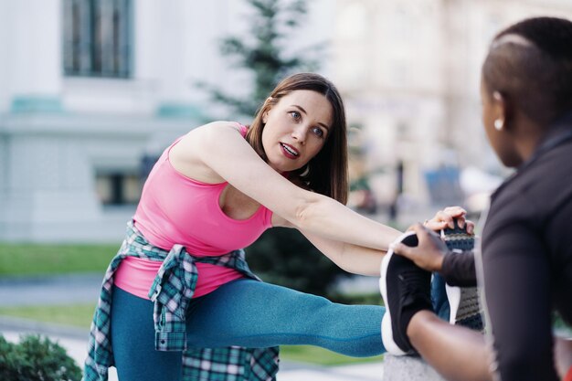 Zwei fitte, vielfältige junge Frauen trainieren gemeinsam auf dem Stadtplatz und machen Dehnübungen und Handverlängerungen