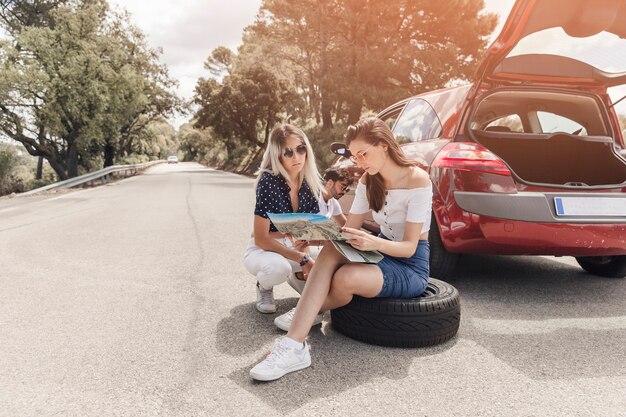 Zwei Frauen, welche die Karte sitzt nahe dem aufgegliederten Auto auf Straße betrachten