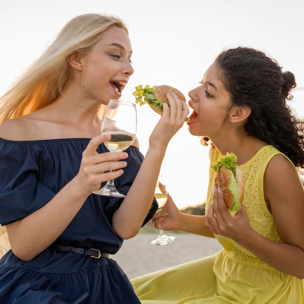 Zwei Freundinnen, die Burger am Strand mit Wein essen