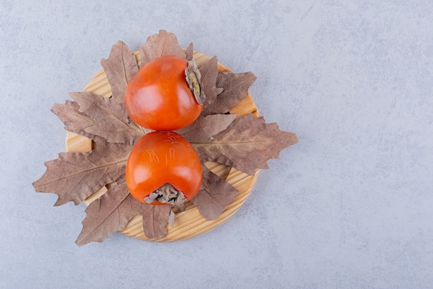 Zwei frische Kakifrüchte und getrocknete Blätter auf Holzplatte.