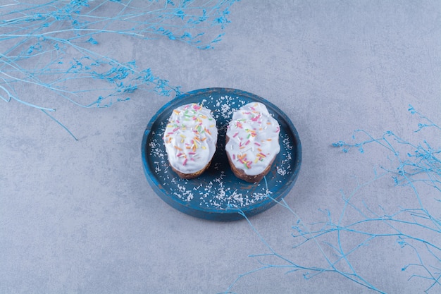 Zwei frische süße Cupcakes mit bunten Streuseln und Sahne auf einem blauen Holzbrett.