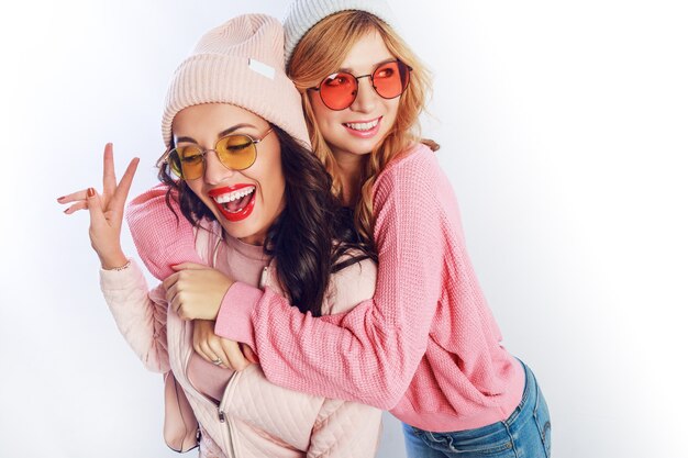 Zwei fröhliche junge Freunde in perfekter Stimmung, die Zeit zusammen auf weißem isoliertem Hintergrund haben. Positive Mädchen tragen rosa kuschelige Pullover und Pullover, trendigen Hut, süße Brille.