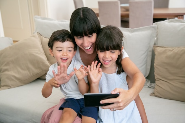 Zwei fröhliche Kinder und ihre glückliche Mutter, die Telefon für Videoanruf benutzen, während sie zusammen auf der Couch zu Hause sitzen