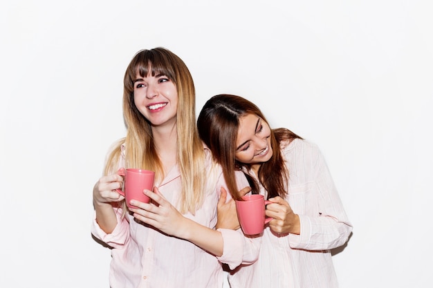 Zwei fröhliche weiße Frauen im rosa Pyjama mit der Tasse Tee, die aufwirft. Flash-Porträt.