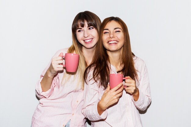 Zwei fröhliche weiße Frauen im rosa Pyjama mit der Tasse Tee, die aufwirft. Flash-Porträt.