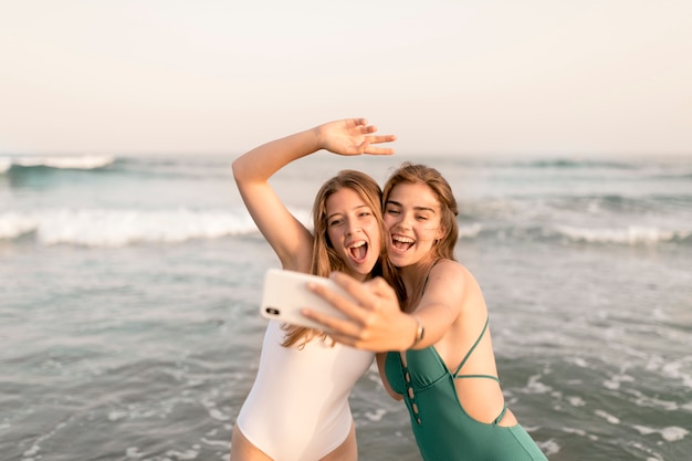 Zwei frohe Freundinnen, die selfie vor Meereswellen nehmen