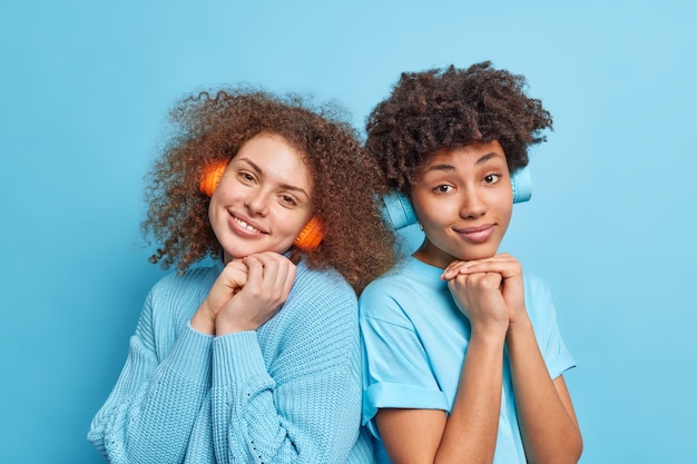 Zwei gemischte beste Freunde, die beiläufig gekleidet sind, stehen eng beieinander und hören Audiospuren in drahtlosen Kopfhörern, verbringen Freizeit zusammen isoliert über blauer Wand. Menschen-Hobby-Konzept