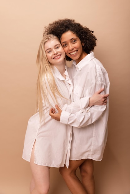 Zwei gemischtrassige junge Mädchen in weißen Hemden stehen in Umarmung und schauen lächelnd in die Kamera auf beigem Hintergrund