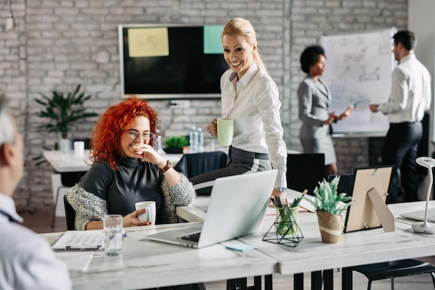 Zwei Geschäftsfrauen lachen, während sie mit ihrem Kollegen sprechen und in einem modernen Büro Kaffee trinken Es gibt Menschen im Hintergrund