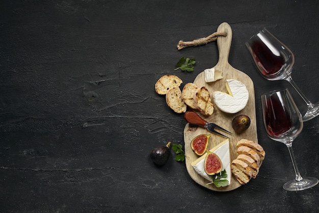 Zwei Gläser Rotwein und eine leckere Käseplatte mit Obst und geröstetem Brot auf einer hölzernen Küchenplatte auf schwarzem Steinhintergrund, Draufsicht, Kopierraum. Gourmetessen und trinken.