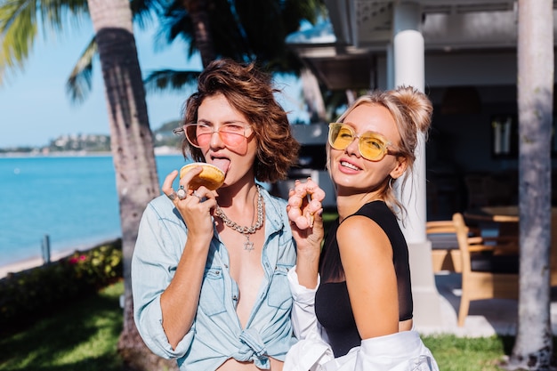 Zwei glückliche fit Frau in der rosa und gelben Sonnenbrille lächelnd, die Spaß lachend mit Donuts, im Freien hat