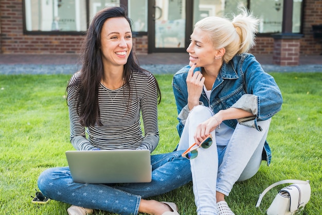 Zwei glückliche Frauen, die auf Gras mit Laptop sitzen