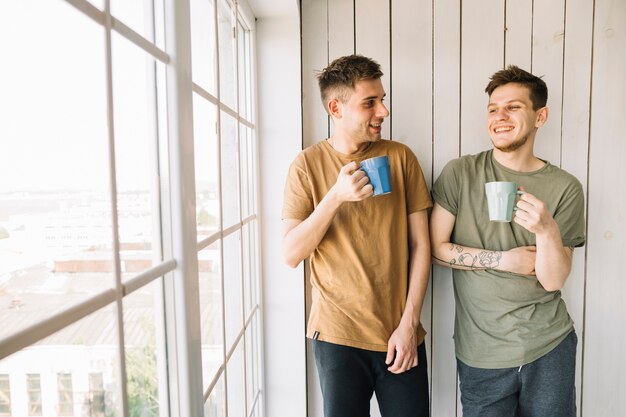 Zwei glückliche Freunde, die den Morgenkaffee steht nahes Fenster trinken