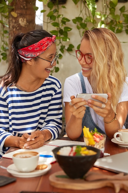 Zwei glückliche Freunde schauen sich ein Video auf dem Handy an, machen nach dem Unterricht eine Kaffeepause, tragen eine Brille, essen köstlichen Salat und trinken Kaffee, posieren vor dem gemütlichen Café-Interieur, das mit dem drahtlosen Internet verbunden ist