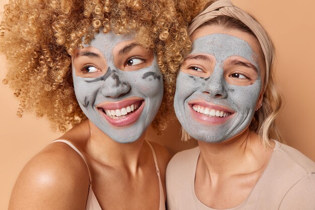 Zwei glückliche, freundliche junge Frau stehen dicht beieinander Appy Gesichtsmasken aus Ton lächeln zahnig konzentriert weg Gesichtsbehandlungen genießen Pflege der Haut Gute Laune Beauty-Konzept