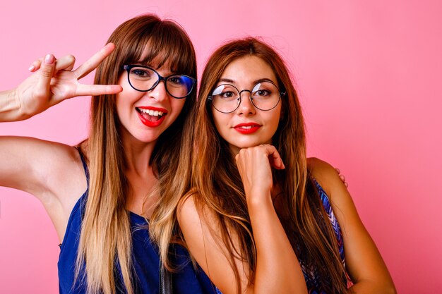 Zwei glückliche fröhliche Frauen lachen und haben Spaß auf der Party, super positive Atmosphäre, glücklich lächelnde Gesichter, beste Hipster-Freunde zusammen, rosa Wand.