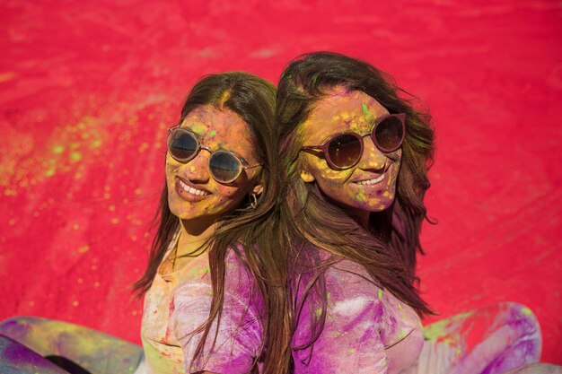 Zwei glückliche junge Frauen bedeckt mit holi Farbe, die Rücken an Rücken sitzt