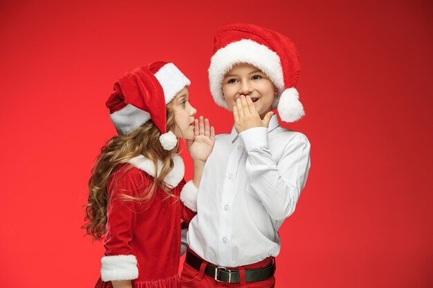 Zwei glückliche Jungen und Mädchen in Weihnachtsmann-Hüten mit Geschenkboxen im roten Studio