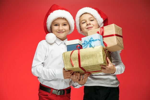 Zwei glückliche Jungs in Weihnachtsmannmützen mit Geschenkboxen auf Rot