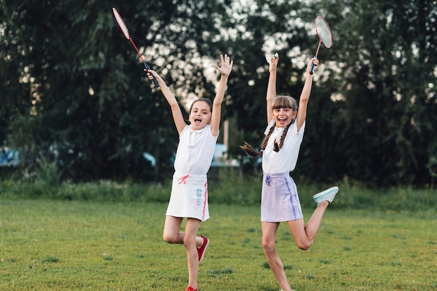 Zwei glückliche Mädchen, welche das Badminton springend in den Park mit Freude halten