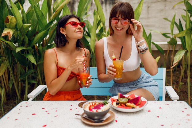 Zwei hübsche Freunde, stilvolle Mädchen, die Freizeit tropisches Café verbringen, leckeres Essen, Smoothie-Schalen, Limonade und Obstteller genießen. Party- und Urlaubsstimmung. Trendige Sonnenbrille.