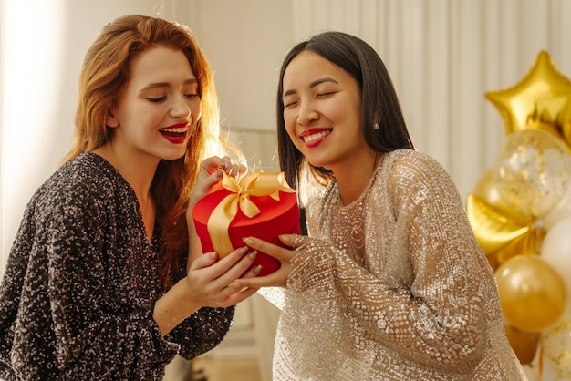 Zwei hübsche junge interrassische Damen in Kleidern packen fröhlich die rote Geschenkbox im Innenbereich aus Überraschungskonzept