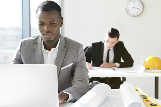 Zwei junge Architekten eines Ingenieurbüros, die im Büro arbeiten. Afrikanischer Designer, der neues Bauprojekt unter Verwendung des Laptops entwickelt und mit Rollen und Lineal am Schreibtisch sitzt.