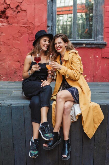 Zwei junge, attraktive, fröhliche Frauen mit Cocktails in den Händen, während sie fröhlich Zeit miteinander im alten, gemütlichen Innenhof des Cafés verbringen