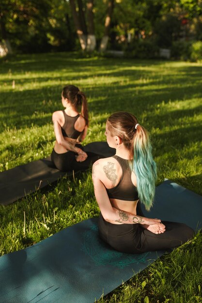 Zwei junge Damen in schwarzen sportlichen Oberteilen und Leggings, die von hinten sitzen und gemeinsam Yoga-Posen trainieren Junge Frauen, die Yoga im Freien praktizieren