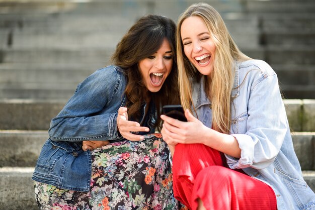 Zwei junge Frauen, die draußen etwas lustige Sache an ihrem intelligenten Telefon betrachten