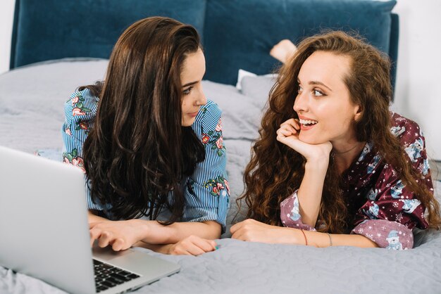 Zwei junge Frauen, die einander mit Laptop auf Bett betrachten