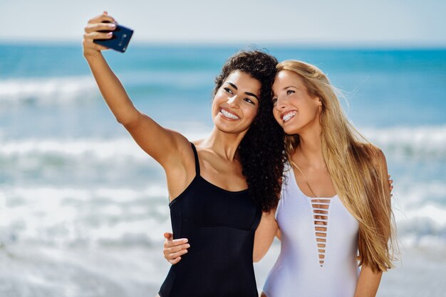Zwei junge Frauen, die selfie mit intelligentem Telefon in den Badeanzügen auf einem tropischen Strand nehmen.