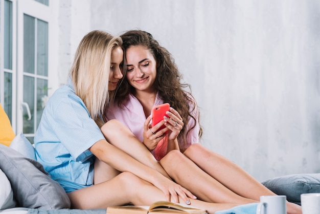 Zwei junge Freundinnen, die auf Bett unter Verwendung des Handys sitzen