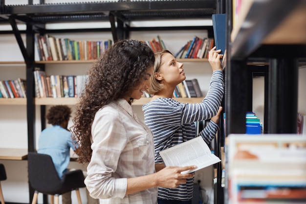 Zwei junge fröhliche Studentinnen in Freizeitkleidung, die in der Nähe von Bücherregalen in der Universitätsbibliothek stehen und Literatur für Teamprojekt durchsehen