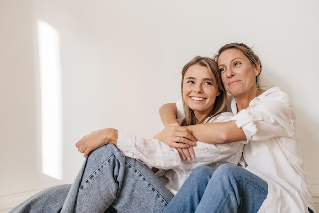 Zwei junge kaukasische frauen tragen lässige kleidung, sitzen auf dem boden vor einer weißen wand und genießen es, zeit miteinander zu verbringen. Freizeit, Lifestyle-Konzept