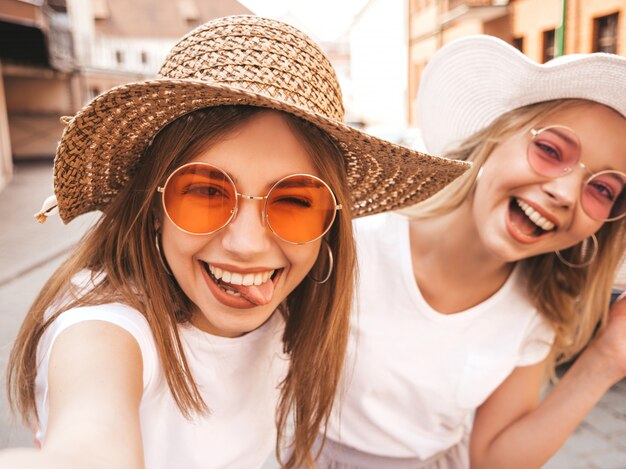 Zwei junge lächelnde blonde Frauen des Hippies im Sommerweißt-shirt. Mädchen, die selfie Selbstporträtfotos auf Smartphone machen Modelle, die auf Straßenhintergrund aufwerfen Frau zeigt Zunge und positive Gefühle