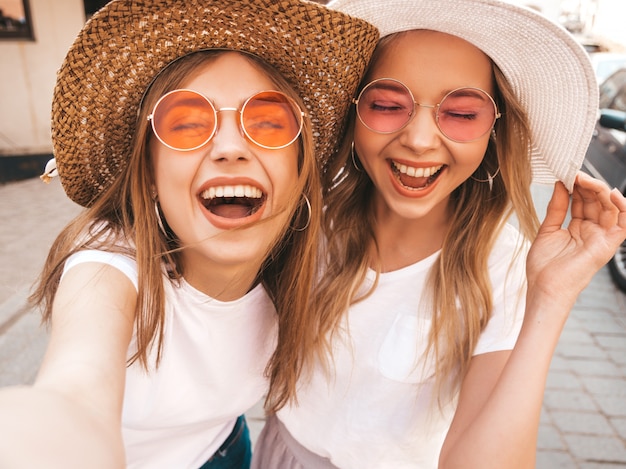 Kostenloses Foto zwei junge lächelnde blonde frauen des hippies im sommerweißt-shirt. mädchen, die selfie selbstporträtfotos auf smartphone machen modelle, die auf straßenhintergrund aufwerfen frau zeigt zunge und positive gefühle