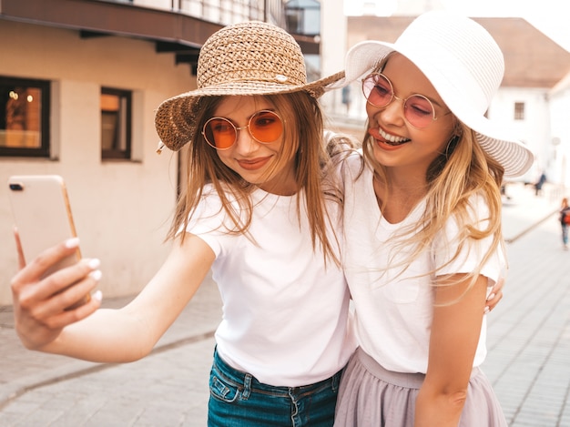Zwei junge lächelnde blonde Frauen des Hippies im weißen T-Shirt des Sommers kleidet. Mädchen, die selfie Selbstporträtfotos auf Smartphone machen.
