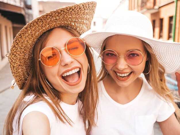 Zwei junge lächelnde blonde Frauen des Hippies im weißen T-Shirt des Sommers kleidet. Mädchen, die selfie Selbstporträtfotos auf Smartphone machen.