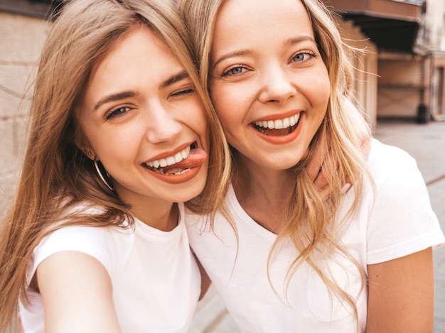 Zwei junge lächelnde blonde Frauen des Hippies im weißen T-Shirt des Sommers kleidet. Mädchen, die selfie Selbstporträtfotos auf Smartphone machen.