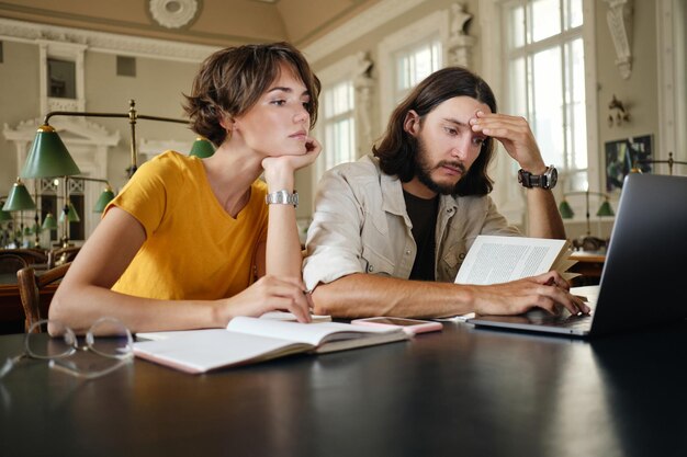 Zwei junge, lockere, nachdenkliche Studenten, die nachdenklich an einem Studienprojekt mit Laptop und Büchern in der Bibliothek der Universität arbeiten