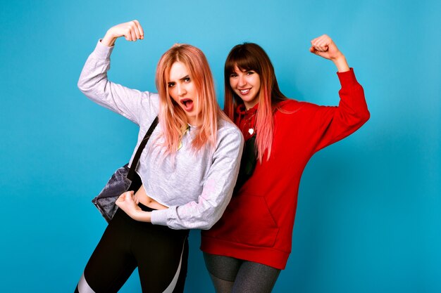 Zwei junge lustige hübsche Hipster-Frauen, die sportliche helle lässige Outfits tragen, Bizeps zeigen und Grimassen machen, zusammen verrückt werden, blaue Wand