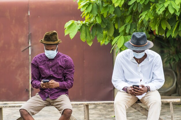 Zwei junge Männer in schützenden Gesichtsmasken, die ihre Telefone benutzen und im Freien sitzen