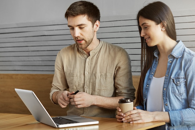 Zwei junge Perspektive Startup-Enthusiasten, die im Café sitzen, Kaffee trinken, über Arbeit sprechen und Projektdetails auf Laptop-Computer durchsehen. Entspannende und produktive Zeit