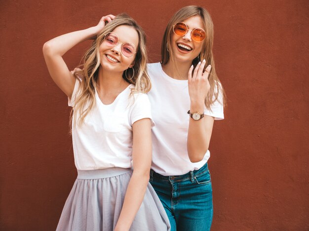 Zwei junge schöne blonde lächelnde Hippie-Mädchen im weißen T-Shirt des modischen Sommers kleidet. Frauen, die in der Straße nahe roter Wand aufwerfen. Positive Models, die Spaß an Sonnenbrillen haben