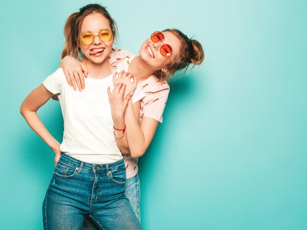 Zwei junge schöne blonde lächelnde Hipster-Mädchen in trendigen Sommer-Hipster-Jeans-Kleidern. Sexy sorglose Frauen, die nahe blauer Wand aufwerfen. Trendige und positive Models, die Spaß an Sonnenbrillen haben