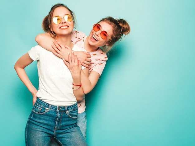 Zwei junge schöne blonde lächelnde hipster-mädchen in trendigen sommer-hipster-jeans-kleidern. sexy sorglose frauen, die nahe blauer wand aufwerfen. trendige und positive models, die spaß haben