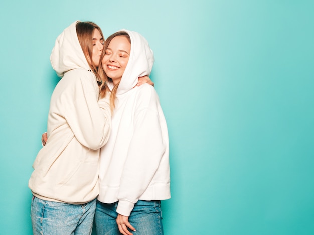 Zwei junge schöne blonde lächelnde Hipster-Mädchen in trendigen Sommer-Hoodie-Kleidern. Sexy sorglose Frauen, die nahe blauer Wand aufwerfen. Model küsst ihre Freundin im Kopf